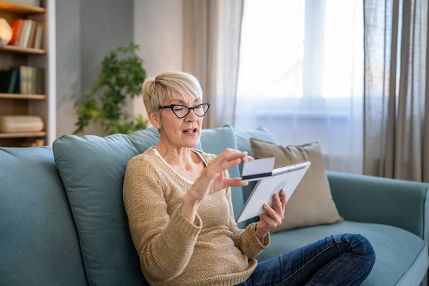 Μια ώριμη ηλικιωμένη γιαγιά κάθεται στο σπίτι χρήση πιστωτικής ή χρεωστικής κάρτας για online αγορές περιηγηθείτε στο διαδίκτυο καταστήματα αγοράζουν πράγματα χρησιμοποιούν ψηφιακό tablet πραγματικό άνθρωποι αντιγράψετε χώρο - Φωτογραφία, εικόνα