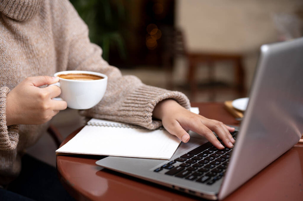 L'immagine ravvicinata di una donna in un accogliente maglione a maglia sta lavorando a distanza in un caffè, sorseggiando caffè caldo mentre lavora sul suo computer portatile. Stile di vita concetto - Foto, immagini