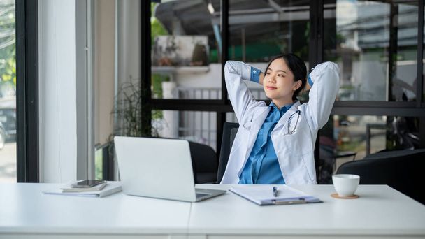 リラックスしたケアフリーのアジアの女性医師が彼女の頭の後ろに手を置き,外を見ながら,彼女の机で仕事の後に休憩しながら夢想的で新鮮な感じをしています. - 写真・画像
