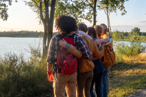 Cette photo capture une vue arrière pleine longueur de cinq jeunes amis multiraciaux du millénaire alors qu'ils célèbrent leur arrivée à un sommet près de la rive d'un lac forestier. Baigné dans la lueur du soleil couchant - Photo, image