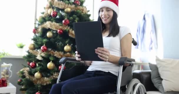 Gehandicapte vrouw in rolstoel zwaaien hand in digitale tablet in de buurt van nieuwjaar boom 4k film slow motion. Gehandicapte kerst op afstand feest concept - Video