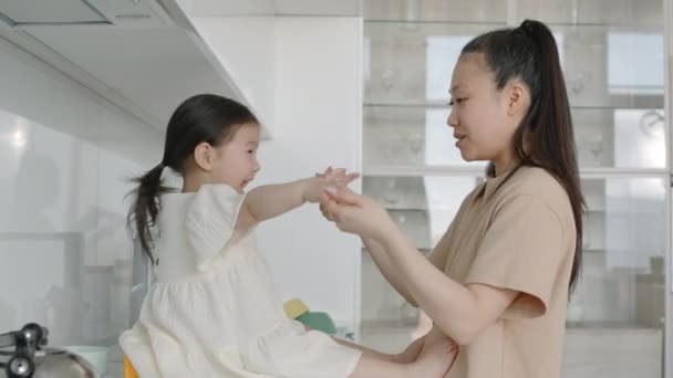 Mediana toma de la joven mujer asiática y su bonita hija pequeña divirtiéndose mientras juegan juego de manos juntos en cocina blanca minimalista durante el tiempo libre - Metraje, vídeo