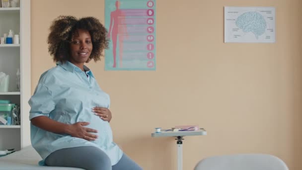 Pan retrato medio de la joven hermosa mujer embarazada negro acariciando su vientre mientras se sienta en el sofá médico en la oficina del ginecólogo y sonriendo a la cámara - Imágenes, Vídeo