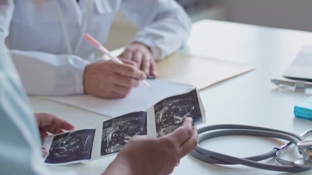 Обрізаний знімок невизначеної вагітної жінки та гінеколога, який сидить за столом у лікарняному кабінеті та обговорює ультразвукове зображення дитини - Кадри, відео