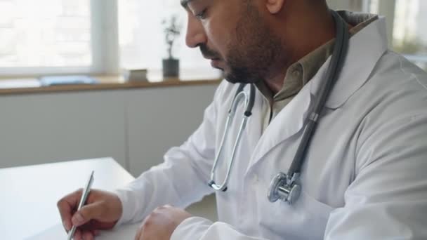 Döntse le a közel-keleti férfi orvos kitöltése orvosi dokumentumokat ül fehér íróasztal ablaka a modern irodában - Felvétel, videó