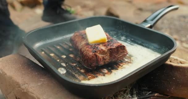 Přidávání másla na steak na pánvi v procesu grilování masa venku. Vaření masa na otevřeném ohni v přírodě na turistické výletě. - Záběry, video