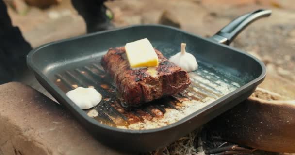 Ajouter du beurre à un steak sur une poêle chaude pendant le grillage de la viande à l'extérieur. Cuisiner de la viande sur un feu ouvert dans la nature lors d'un voyage touristique. - Séquence, vidéo