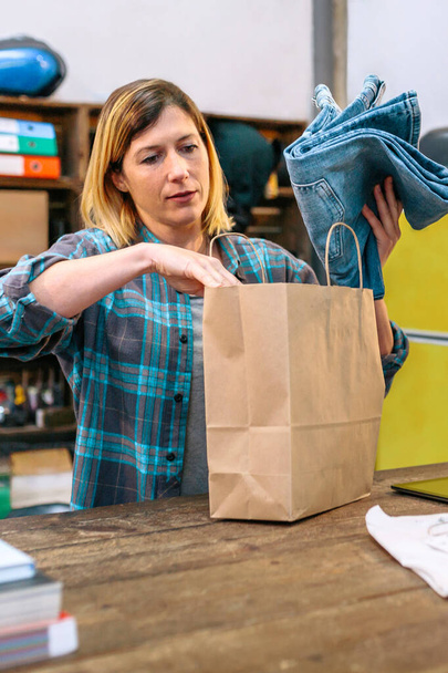 Concentrated woman shop assistant putting jeans into a ecologic kraft paper bag in local store. Female employee preparing clothing to online shop customer. Parcel delivery and ecommerce concept. - Photo, Image