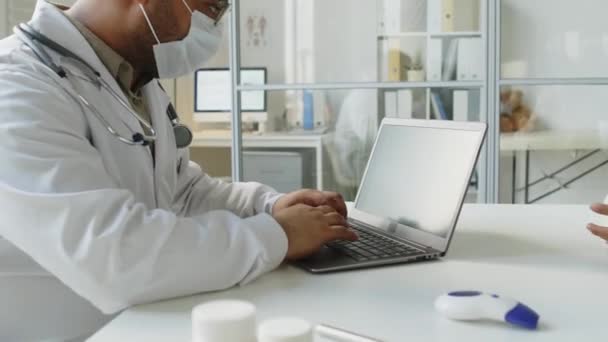 Περιμένετε μέχρι τα μέσα της ενήλικης Μέσης Ανατολής αρσενικό γιατρό φορώντας λευκή ρόμπα εργαστηρίου και μάσκα προσώπου κρατώντας σημειώσεις στο laptop, ενώ ακούτε αγνώριστη ασθενή κατά τη διάρκεια του check up ραντεβού - Πλάνα, βίντεο