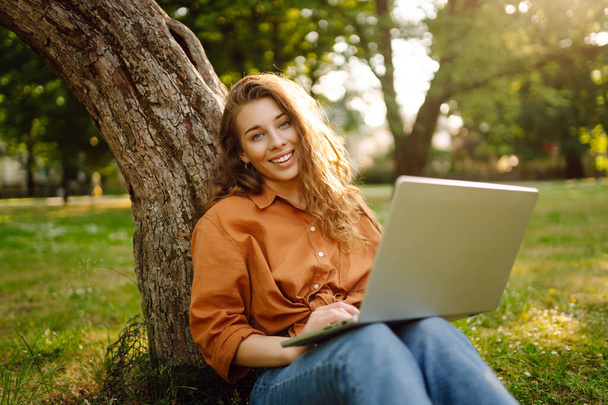 Happy woman with curly hair with a laptop on a green meadow at sunset. Young freelancer woman working outdoors. Freelance work, nature concept. - Foto, imagen