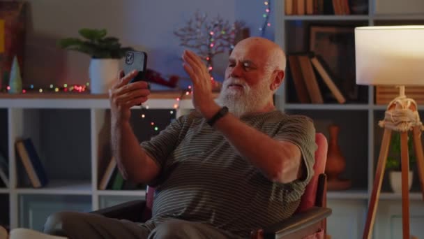 Un uomo anziano in buona salute e di buon umore si siede in camera su una poltrona, utilizza uno smartphone e una videochiamata conversazione. Una mano di onde umane come segno di saluto, sorride, parla animatamente e - Filmati, video