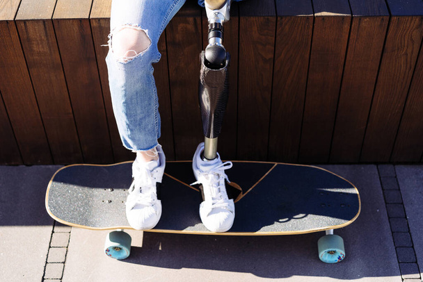 Close up of women prosthetic leg on skateboard in city. Disabled woman with prosthetic leg sitting on wood bench. Woman with leg prosthesis equipment. - Photo, Image