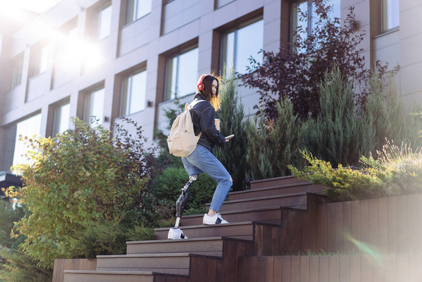 Молодая студентка с протезной ногой поднимается по городу. Женщина-инвалид с протезной ногой по лестнице. Женщина с протезированием ног. - Фото, изображение