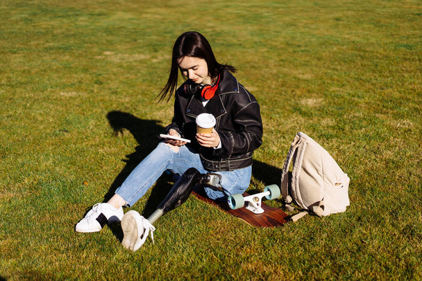 Young woman student with prosthetic leg sitting on green grass in university campus. Disabled woman with bionic leg. Woman with leg prosthesis equipment using phone. - Photo, Image