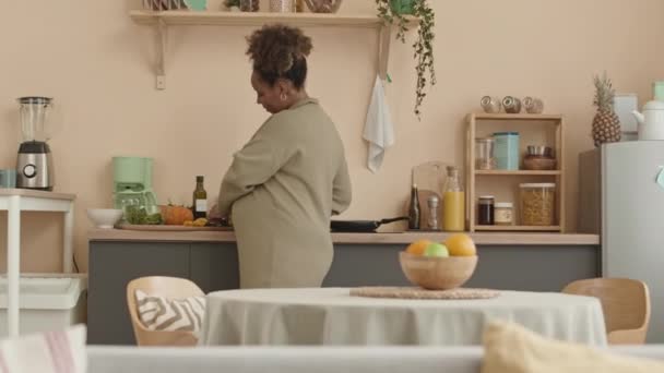 Közepes lövés a fiatal pozitív terhes fekete nő bézs kötött ruha eszik uborka szeletek, miközben az egészséges növényi saláta ebédre hangulatos lakás - Felvétel, videó