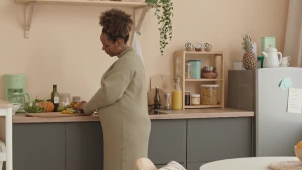 Medium shot of healthy young pregnant African American woman snacking with fresh cucumber slices while standing at cozy kitchen in lunchtime - Footage, Video