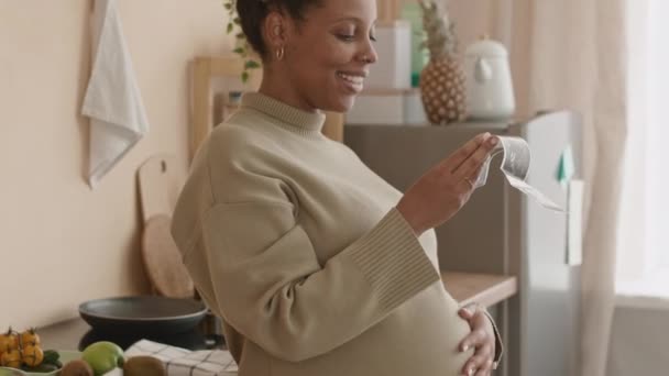 Tilt πλάνο της νεαρής εγκύου Μαύρη γυναίκα χαμογελά και χαϊδεύοντας την κοιλιά της, ενώ κοιτάζοντας υπερηχογράφημα μωρό σάρωση, στέκεται σε ζεστή κουζίνα κατά τη διάρκεια της ημέρας - Πλάνα, βίντεο