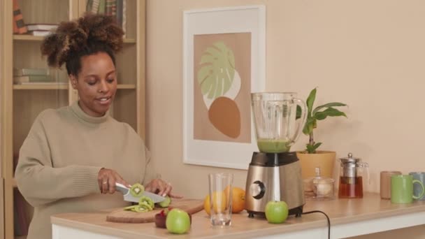Junge schwangere Schwarze schneidet Kiwi und legt Scheiben in den Mixer, während sie zu Hause einen gesunden grünen Smoothie zubereitet - Filmmaterial, Video