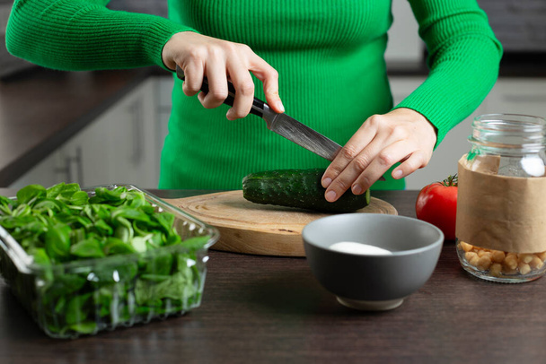 Vrouw snijdt komkommer voor het koken van groentesalade. Huiskeuken. - Foto, afbeelding