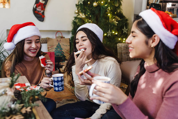 grupo de amigos latinos comiendo galletas y chocolate caliente sentados cerca del árbol de Navidad en casa en México. Fiestas y celebraciones en América Latina - Foto, imagen