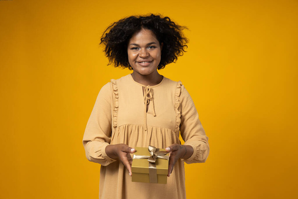Mosolygós göndör nő bézs ruha ajándékdoboz a kezében sárga háttér - Fotó, kép