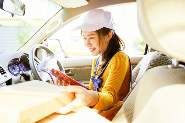 美しいアジアの女性の小包配達サービスドライバーのピックアップは,顧客の小包箱をオンラインビジネスが座って,顧客に送信する前に正確なレコードクリップボードをチェックします. - 写真・画像