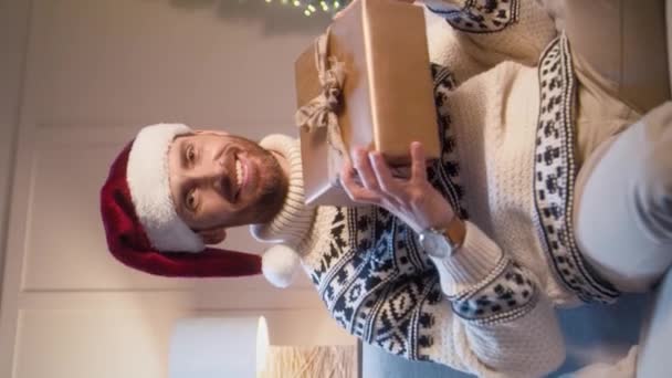 Hombre caucásico guapo con sombrero rojo de Santa Claus disfrutando de regalo para las vacaciones de Navidad. Caja de espera masculina barbuda con presente mientras está sentado en el sofá. Sonriendo alegremente con humor festivo. - Metraje, vídeo