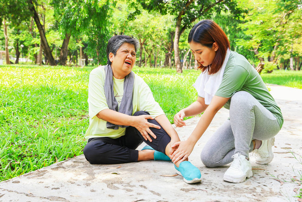 Oudere vrouw zit grond pijn in haar linkerenkel tijdens het sporten joggen in de tuin het ontvangen van eerste hulp van haar dochter voor verstuikte enkel pijn met voorzichtigheid: Oudere gezondheid in de verzekering. - Foto, afbeelding