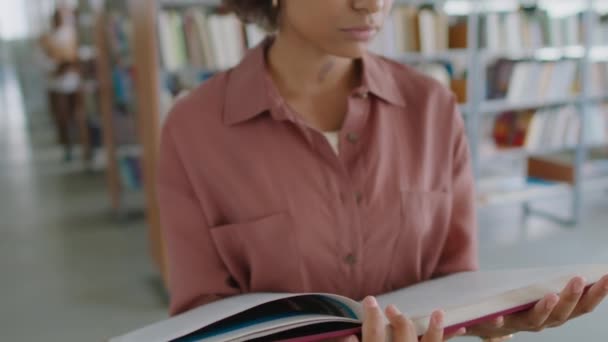 Inclinado hacia arriba retrato de alegre estudiante afroamericana con grueso diccionario libro en las manos sonriendo a la cámara en la biblioteca moderna - Metraje, vídeo