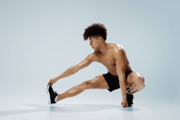 Posilovací cvičení. Mladý atletický muž bez košile cvičení v prodloužené nohy squat póza, protahování nohou a dotýkání prstů, demonstrující flexibilitu a sílu nad šedým studiem pozadí - Fotografie, Obrázek