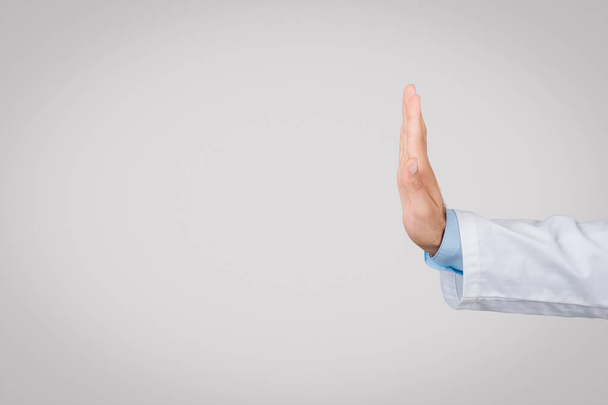 Joven médico europeo en bata blanca, mostrando gesto de mano parada, vista lateral, lo que significa precaución o advertencia, sobre fondo gris simple, espacio libre - Foto, imagen