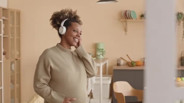 Střední záběr radostné mladé černošky těhotná žena tanec na hudbu v bezdrátových sluchátkách v útulném domě - Záběry, video