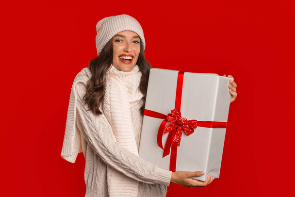 Cheerful millennial lady wearing Santa hat holds wrapped present box with anticipation of Christmas on red studio background. Perfect banner for holiday season advertisements - Photo, Image