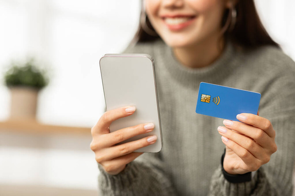 Elinde telefon ve mavi plastik banka kredi kartı olan ev içini tutan genç bir kadın. Kadın alışverişi ya da internet bankacılığı, internet hizmetleri - Fotoğraf, Görsel