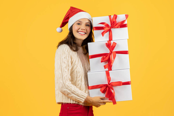 Señora feliz sosteniendo múltiples cajas de Navidad pila, posando alegre y festivo en el sombrero de Santa, perfecto para las compras de vacaciones y temas de celebración, estudio de tiro sobre fondo amarillo - Foto, imagen
