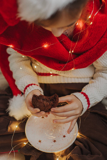 Hands of a teenage girl holding a cookie against the background of a Santa hat and garland lights. Festive New Year food concept. Close-up photo. - Photo, Image