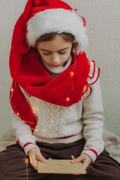 Uma menina adolescente bonito em um pulôver e chapéu de Papai Noel segurando um envelope com uma carta para Papai Noel contra o fundo de uma parede de luz. Ambiente de Natal mágico em casa. - Foto, Imagem