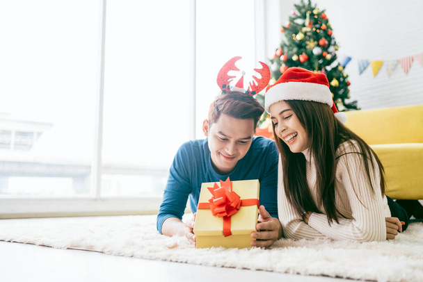 Joven mujer asiática feliz usando un sombrero de Santa Claus con su novio con un regalo de Navidad mientras se acuesta en la alfombra con un árbol de Navidad en el fondo. Foto con espacio de copia. - Foto, imagen