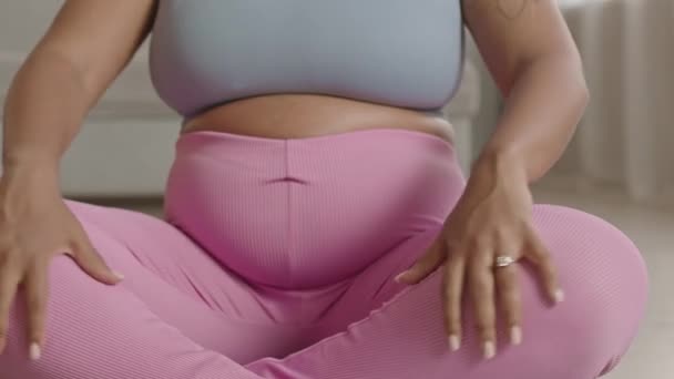 Colpo ritagliato di donna incinta irriconoscibile che indossa stretti leggings rosa seduti in posa di loto sul tappeto yoga a casa e meditando - Filmati, video