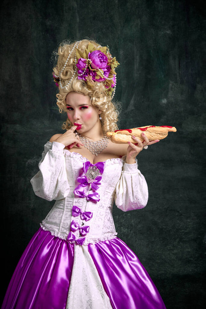 Nainen pukeutunut kuin klassinen keskiaikainen henkilö, kuningatar syö kuuma, herkullinen ja valtava amerikkalainen hot dog vastaan vintage tausta. Ruoka, ravitsemus, taide, ateriapalvelu, ravintolavalikko, toimitus. - Valokuva, kuva