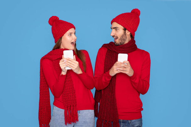 Oferta de Navidad. Retrato de pareja joven conmocionada en sombreros de invierno sosteniendo teléfonos inteligentes y mirándose entre sí con emoción, hombre y mujer sorprendidos reaccionando emocionalmente a las ventas en línea, espacio de copia - Foto, imagen