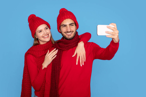 Selfie de invierno. Pareja joven feliz en sombreros de punto haciendo fotos en el teléfono inteligente, Hombre alegre y mujer abrazando mientras posan en la cámara del teléfono móvil, de pie aislado sobre fondo azul - Foto, imagen