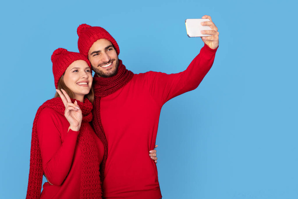 Joyeux jeune couple portant des chapeaux d'hiver s'amuser tout en prenant Selfie sur Smartphone, heureux homme et femme européenne riant et montrant geste de paix à la caméra, posant isolé sur fond bleu - Photo, image