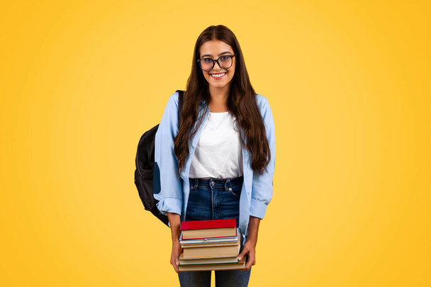 Positive teenager student lady in glasses hold many books, enjoy study and homework, isolated on yellow studio background. Education, lifestyle at school, ad and offer at university - Photo, Image