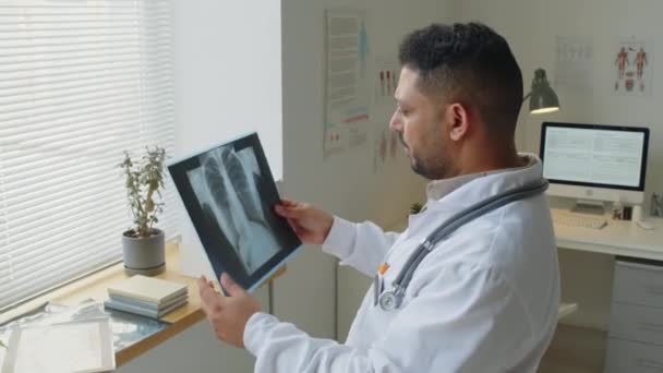 Közepes felvételű közel-keleti férfi orvos fehér köpenyben áll az ablak előtt, miközben a beteg tüdő röntgenjét vizsgálja. - Felvétel, videó