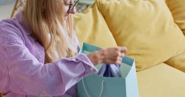 Jonge blonde vrouw maakt winkelen in een moderne winkel. Lady horloges stijlvolle t-shirt genomen uit papieren zak zitten op de bank. Vrouw voelt zich blij met aankoop - Video