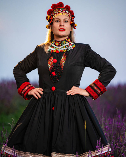 Dívka v chelsea čelence a černé šaty zdobené červenou výšivkou stojí mezi levandulovými keři. - Fotografie, Obrázek