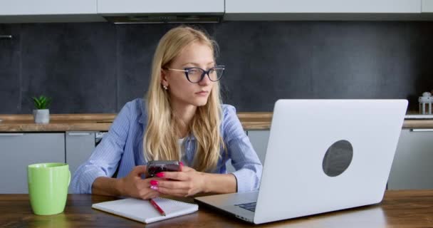 Mujer rubia en gafas busca información en Internet utilizando el ordenador portátil en casa. Mujer joven utiliza el teléfono móvil para estudiar. Comunicación moderna - Imágenes, Vídeo
