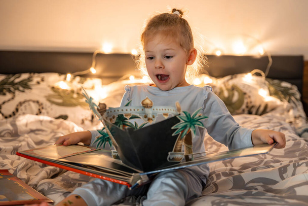 Petite fille lecture pop up livre dans le lit à la maison dans l'environnement de Noël avec fond de lumières. Photo de haute qualité - Photo, image