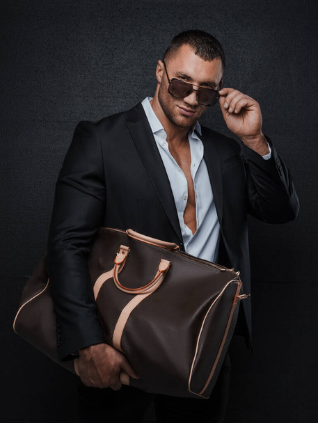 Homem elegante em óculos de sol, em um terno elegante com uma camisa desabotoada, mostrando seu tronco muscular, posando com um luxuoso saco de alta moda em um fundo cinza - Foto, Imagem